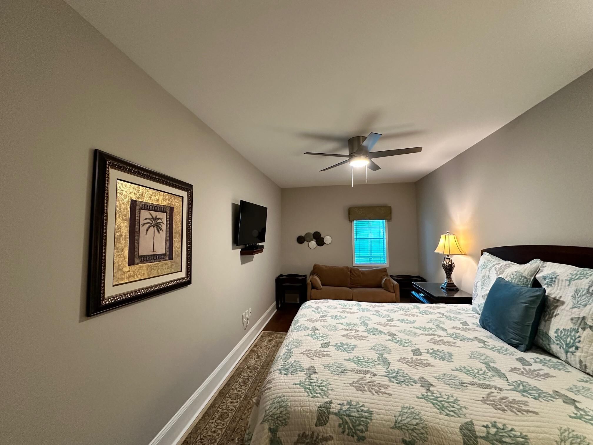 A bedroom with a bed, a ceiling fan, a TV, a window with blinds, and a framed picture on the wall.