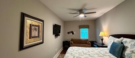 A bedroom with a bed, a ceiling fan, a TV, a window with blinds, and a framed picture on the wall.