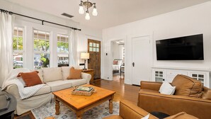 A spacious living room with a sofa, coffee table, and leather armchair. A television is mounted on the wall, and a wooden door leads to another room. - Luxury Paso Bungalow • Hot Tub & EV Charger (Paso Robles)
