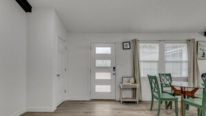 Une pièce avec un plancher en bois, des murs blancs, une porte avec des panneaux de verre dépoli, une petite table avec des chaises et une fenêtre avec des stores.