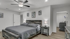 Une chambre à coucher comprenant un lit, une table de chevet, une commode, un ventilateur de plafond et une porte donnant sur une autre pièce.