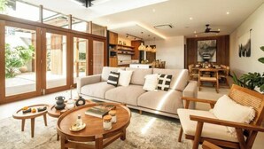 A modern living room with a sofa, coffee table, and wooden chairs. There is a dining area with a table and chairs. The room has large windows and a view of the outdoors. - Big pool 5 BR Villa Canggu Prime location, 5 min to Berawa beach, . (Tibubeneng)