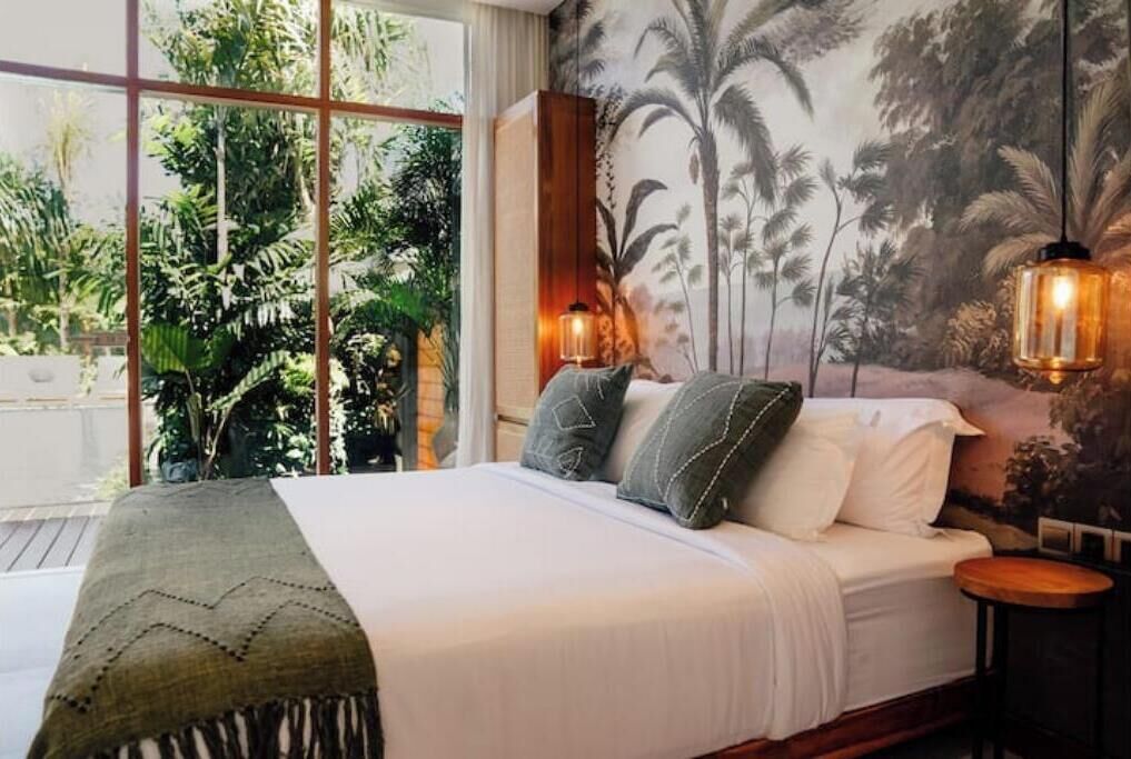 Ein Schlafzimmer mit einem großen Bett, Blick auf Grün durch ein Fenster und einem tropischen Wandbild.