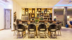 A modern dining area with a central round table, surrounded by six chairs. There is a built-in kitchen with wooden shelves and a refrigerator.