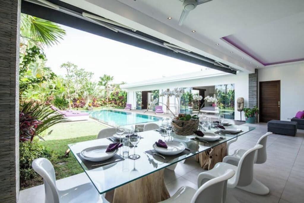 Un espace de salle à manger moderne avec une table en verre, des chaises blanches et une vue sur une piscine et un jardin.