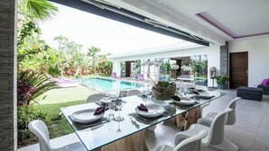 A modern dining area with a glass table, white chairs, and a view of a swimming pool and garden. - Cozy 4 BR Modern Villa in Seminyak, Large Pool, Perfect and Fantastic (Kerobokan Kelod, Kec. Kuta Utara)