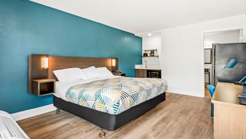 A modern hotel room with a large bed, a bedside table, a nightstand, a desk with a chair, a microwave, and a small kitchenette.