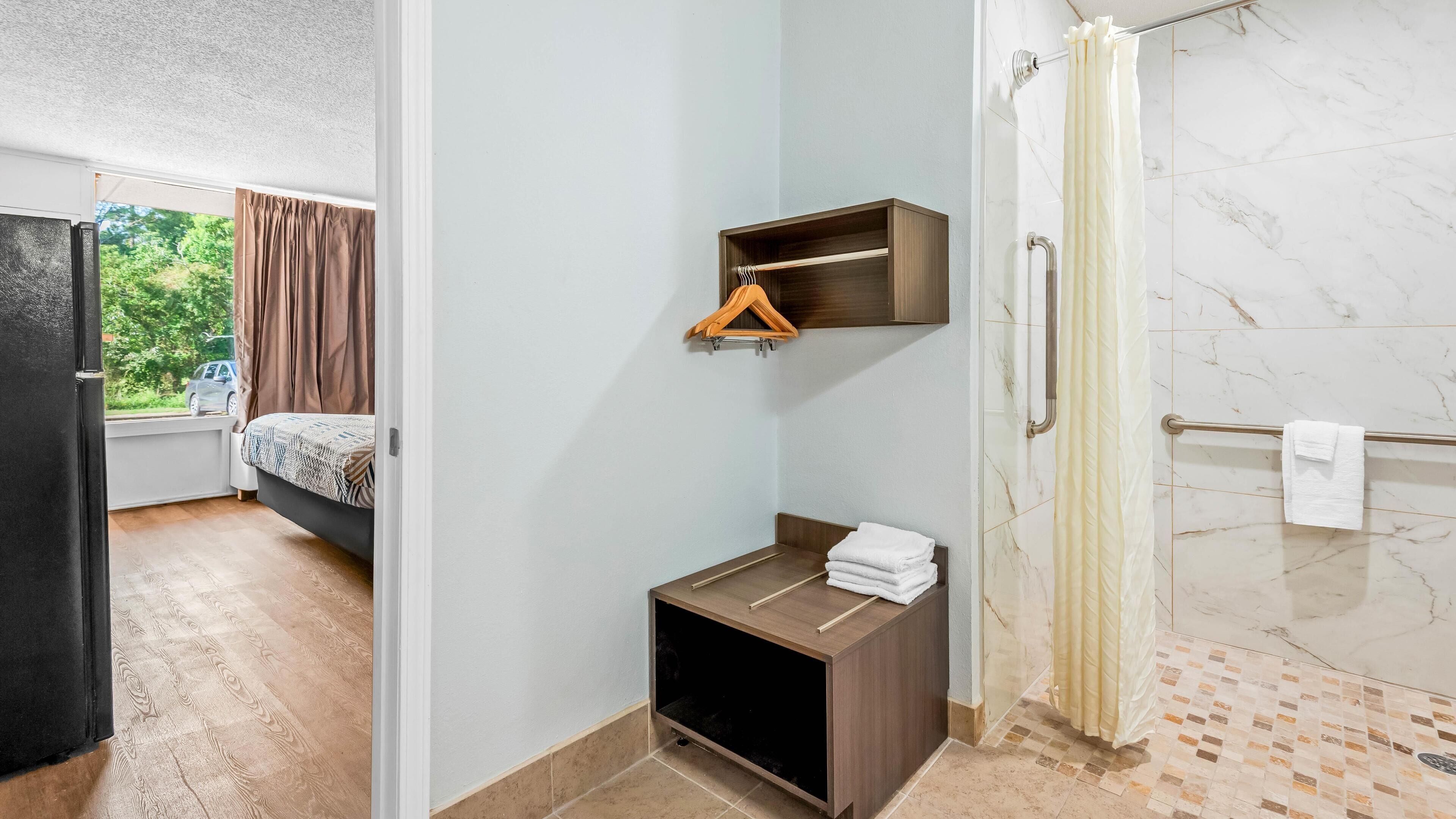 A hotel room with a bed, a small wooden cabinet, a towel rack, and a shower area with a marble floor.