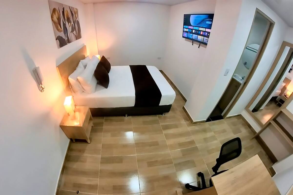 A hotel room with a bed, bedside table, lamp, TV, and a desk with a chair.