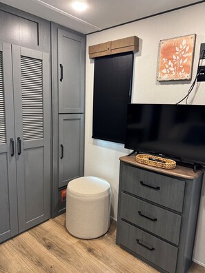 Ein Zimmer mit grauen Schränken, einem Flachbildfernseher auf einem Holzständer, einem kleinen Hocker und einem an der Wand befestigten Regal mit einem gewebten Korb.