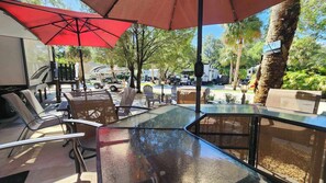 Eine Terrasse im Freien mit Glastischen und -stühlen, roten Sonnenschirmen und Blick auf parkende Fahrzeuge und Bäume.