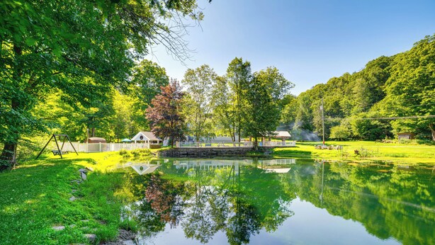 Un paysage paisible au bord d’un lac, avec un reflet limpide dans l’eau, un pont de pierre et un petit bâtiment en arrière-plan.