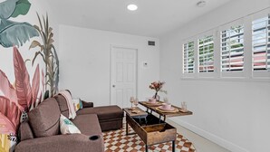 Ein Wohnzimmer mit Schachbrettmuster-Boden, einem braunen Sofa, einem hölzernen Couchtisch und einer Wand mit großformatiger Kunst von tropischen Blättern.