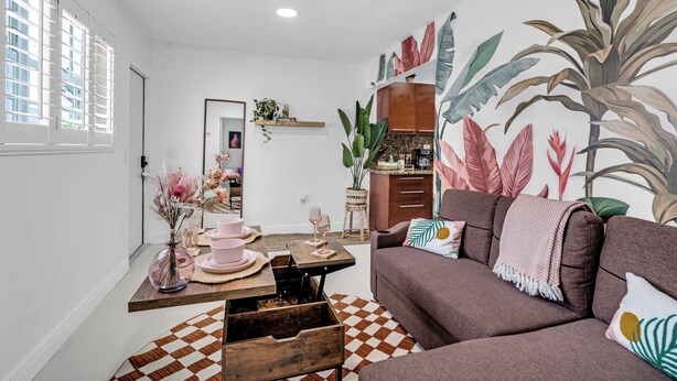 Ein Wohnzimmer mit braunem Sofa, einem Holztisch und einem karierten Teppich. Es gibt eine Küchenzeile mit einem Holzschrank und einer Pflanze auf der Arbeitsplatte.