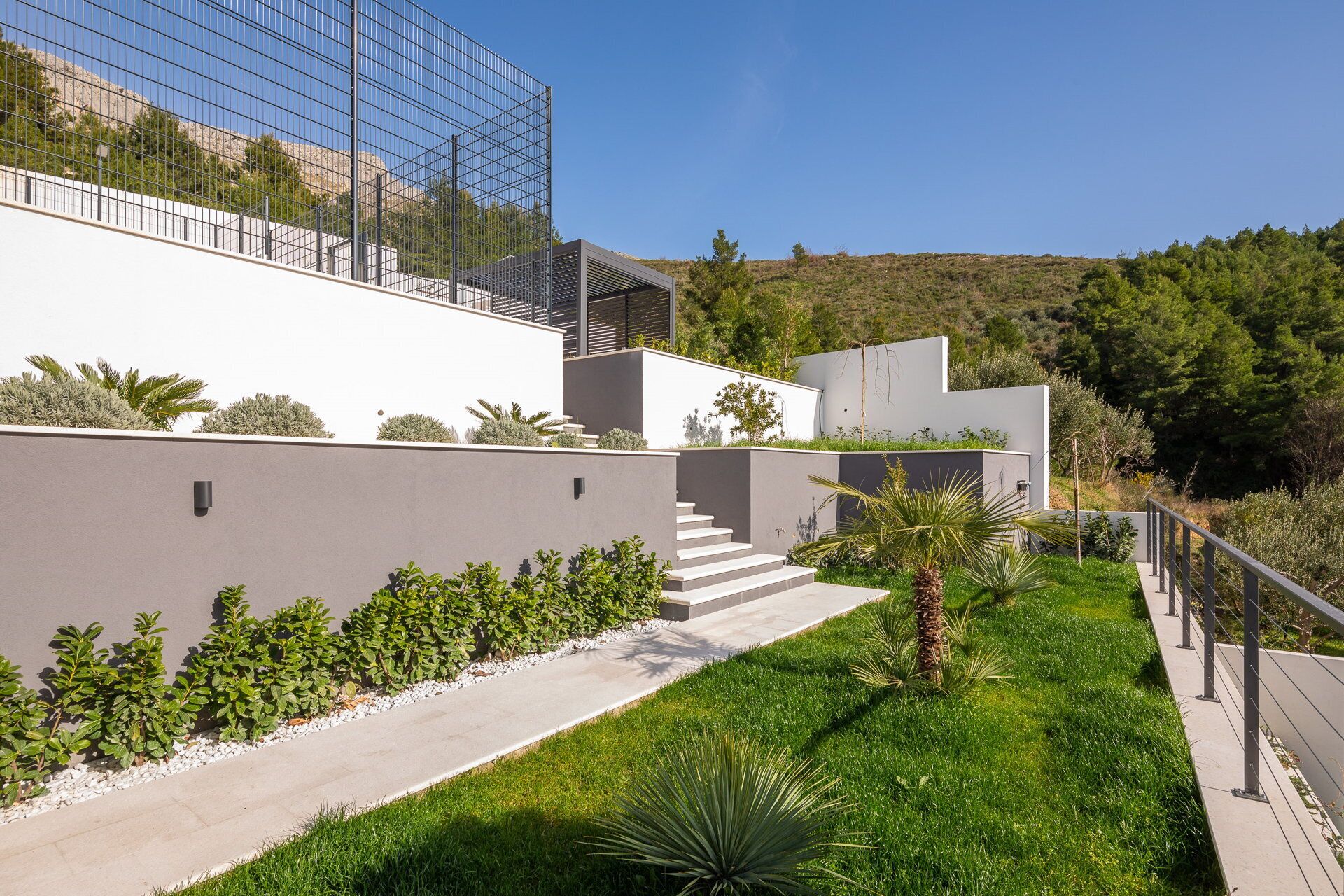 A modern building with a glass facade, a concrete pathway, and a well-maintained garden with various plants and a palm tree.