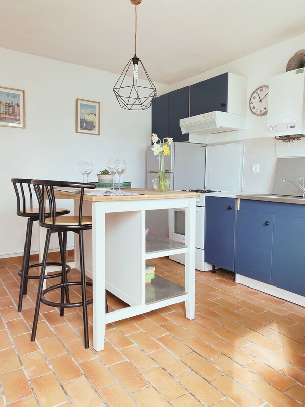 A kitchen with a dining area, featuring a white table, black chairs, a hanging light fixture, and a blue cabinet. - Gite : L'Érable Normand (Thue et Mue)