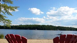 To røde Adirondack-stole med udsigt til en sø, hvor kajakroere ses i det fjerne, og en skovkant løber langs bredden.