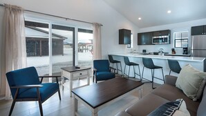 Una cocina moderna con armarios oscuros y electrodomésticos de acero inoxidable, un comedor con sillas azules y una mesa de madera, y un amplio ventanal corredero que da a un área exterior nevada.