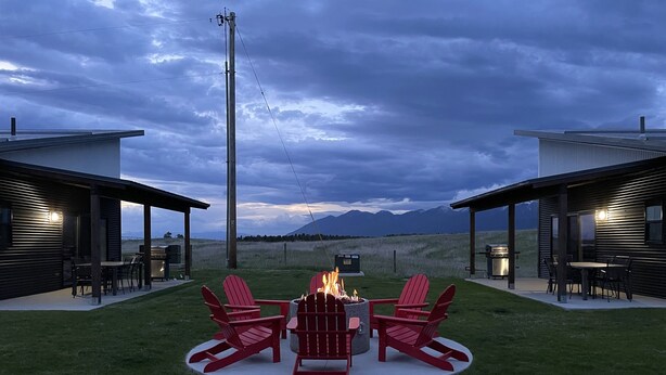 Un área de estar moderna al aire libre con un brasero y sillas Adirondack rojas, rodeada de un césped cuidado y con vistas a un paisaje montañoso.