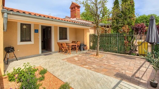 A house with a tiled patio, a dining area with chairs, a barbecue grill, and a fenced garden.