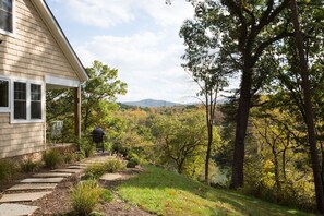 Une maison dotée d’un porche abrité, d’un barbecue et d’un chemin de pierre menant à une zone boisée, avec des arbres et des collines en arrière-plan.