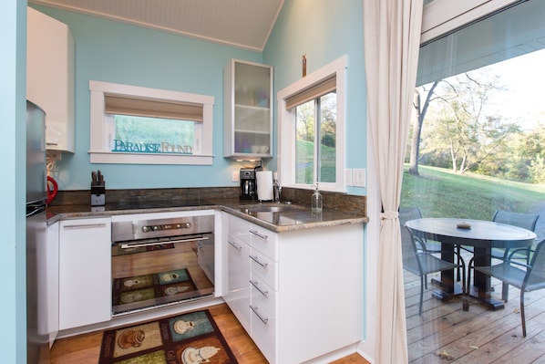 A kitchen with white cabinets, a built-in oven, a window with a view of a green landscape, and a dining area with a table and chairs.