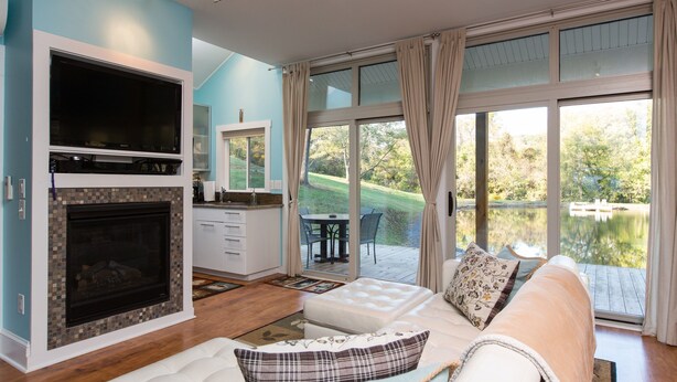 A modern living room with a fireplace, a sofa with decorative pillows, and a large window offering a view of a lake and greenery. - Fantasy Cottage: Private Pond, Waterfall, Hot Tub (Front Royal)