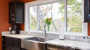 Una cocina con un lavaplatos de acero inoxidable, armarios oscuros, una cafetera y una ventana con vistas a árboles.