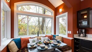 Comedor con una mesa dispuesta para cuatro personas, ventanas amplias y una cocina con armarios oscuros.