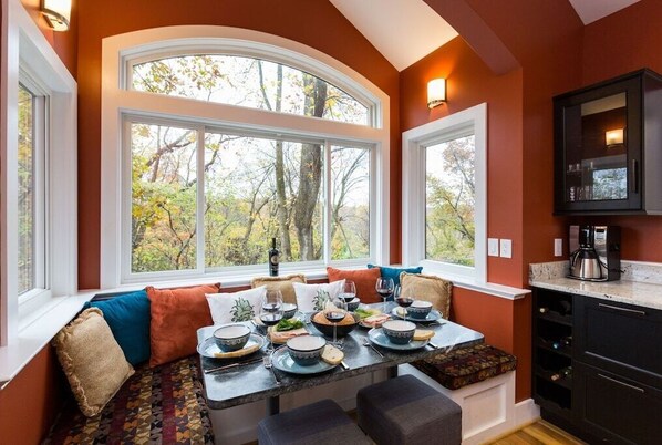 A dining area with a table set for four, large windows, and a kitchen area with dark cabinetry.
