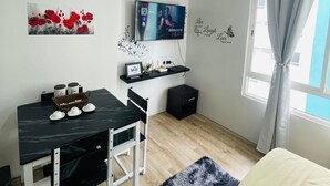 Een moderne, goed verlichte kamer met een eettafel, stoelen, een aan de muur bevestigde televisie en een raam met gordijnen.