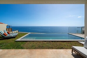 A swimming pool with a view of the ocean, a grassy area with lounge chairs, and a building with a red roof.