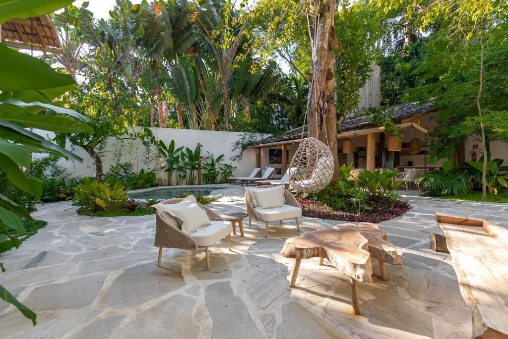 A stone patio with outdoor furniture, a hanging chair, and a pool area surrounded by lush greenery.