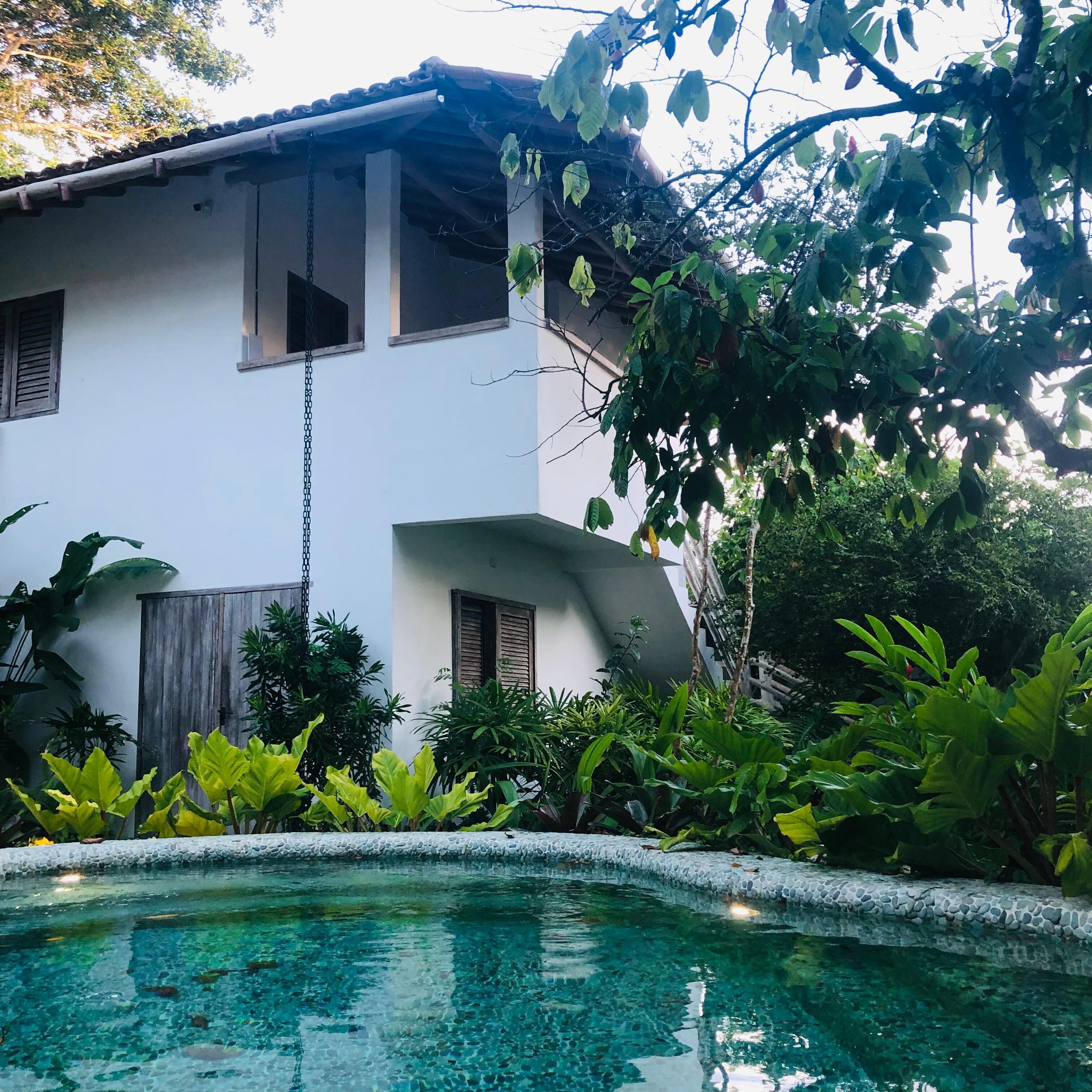 A white building with a tiled roof, surrounded by lush greenery and a clear pool.