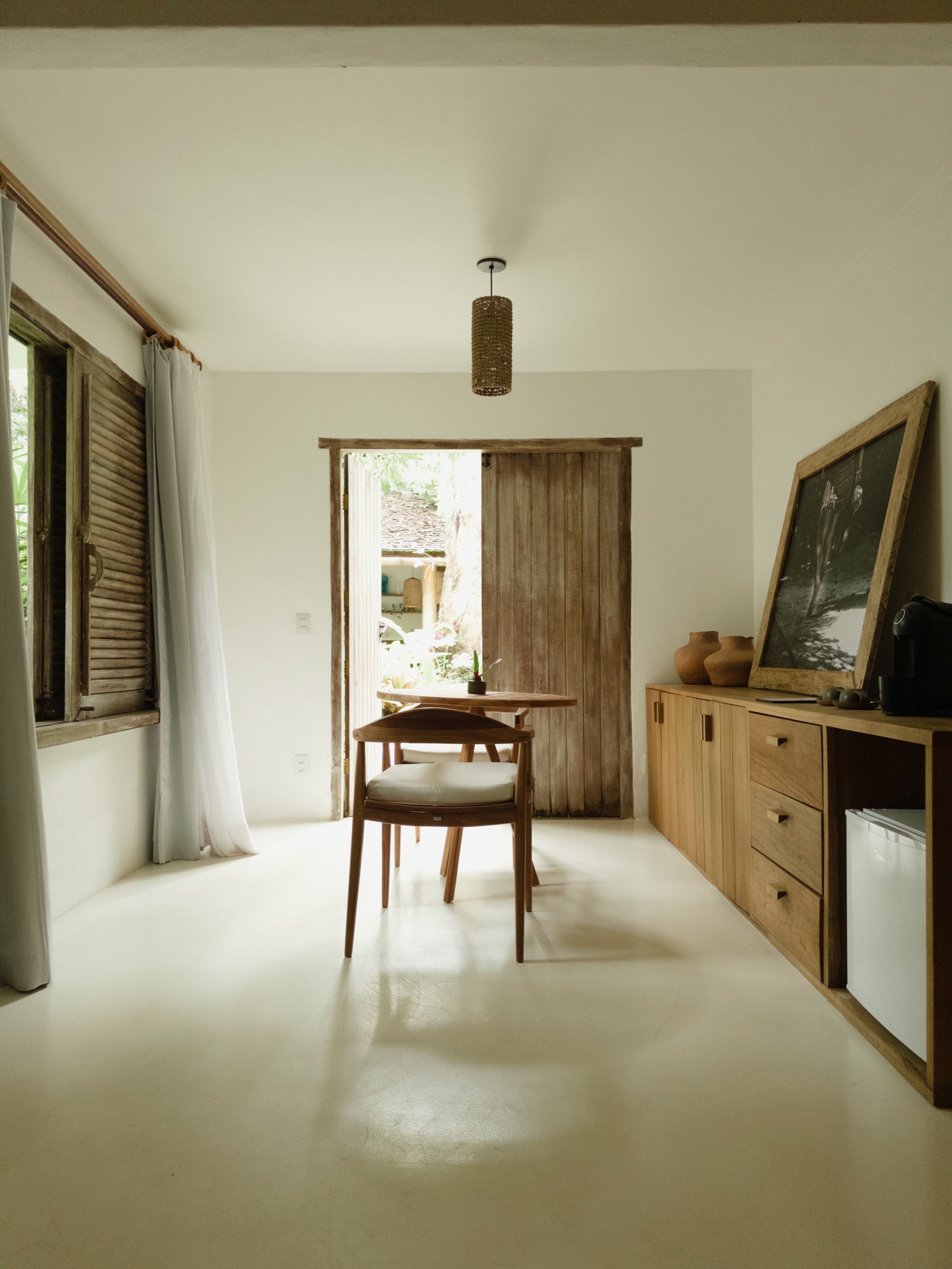 A room with a wooden table and chair, a cabinet with a painting, and a view through a door.