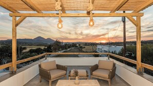 A covered outdoor seating area with wicker furniture and a table, overlooking a scenic landscape with mountains and a sunset.
