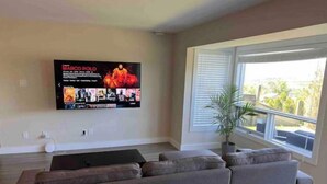 A modern living room with a large flat-screen TV mounted on the wall, a grey sectional sofa, a small coffee table, and a large window with blinds. - Private 1bd 1 ba suite w/ lake views (Vernon)