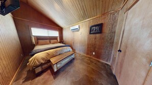 Une chambre avec un plafond en bois, un lit avec une banquette, une fenêtre avec des stores, un climatiseur et un tableau encadré au mur.