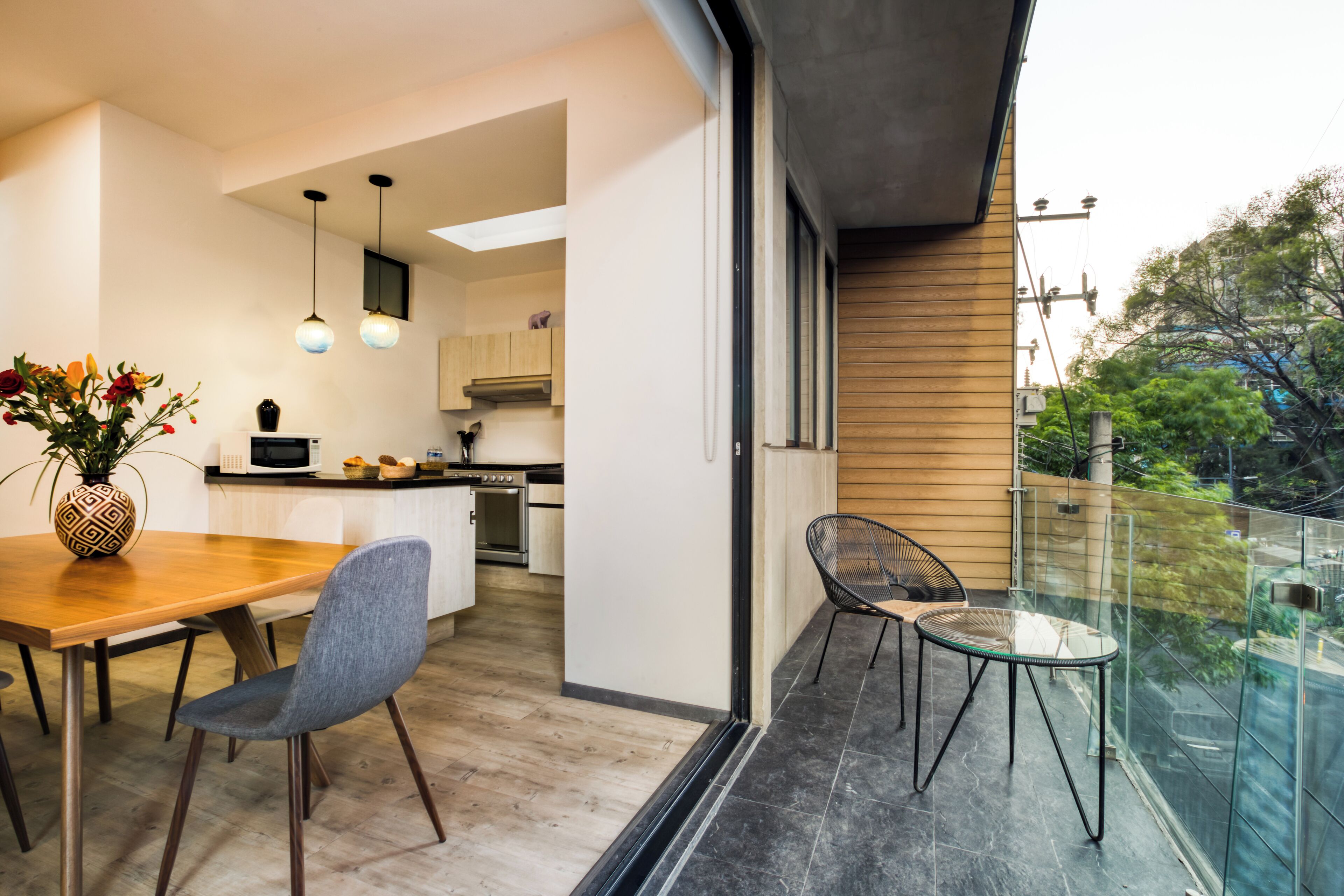 Una cocina moderna con armarios de madera, un comedor con mesa y sillas de madera, y un balcón con barandilla de vidrio.