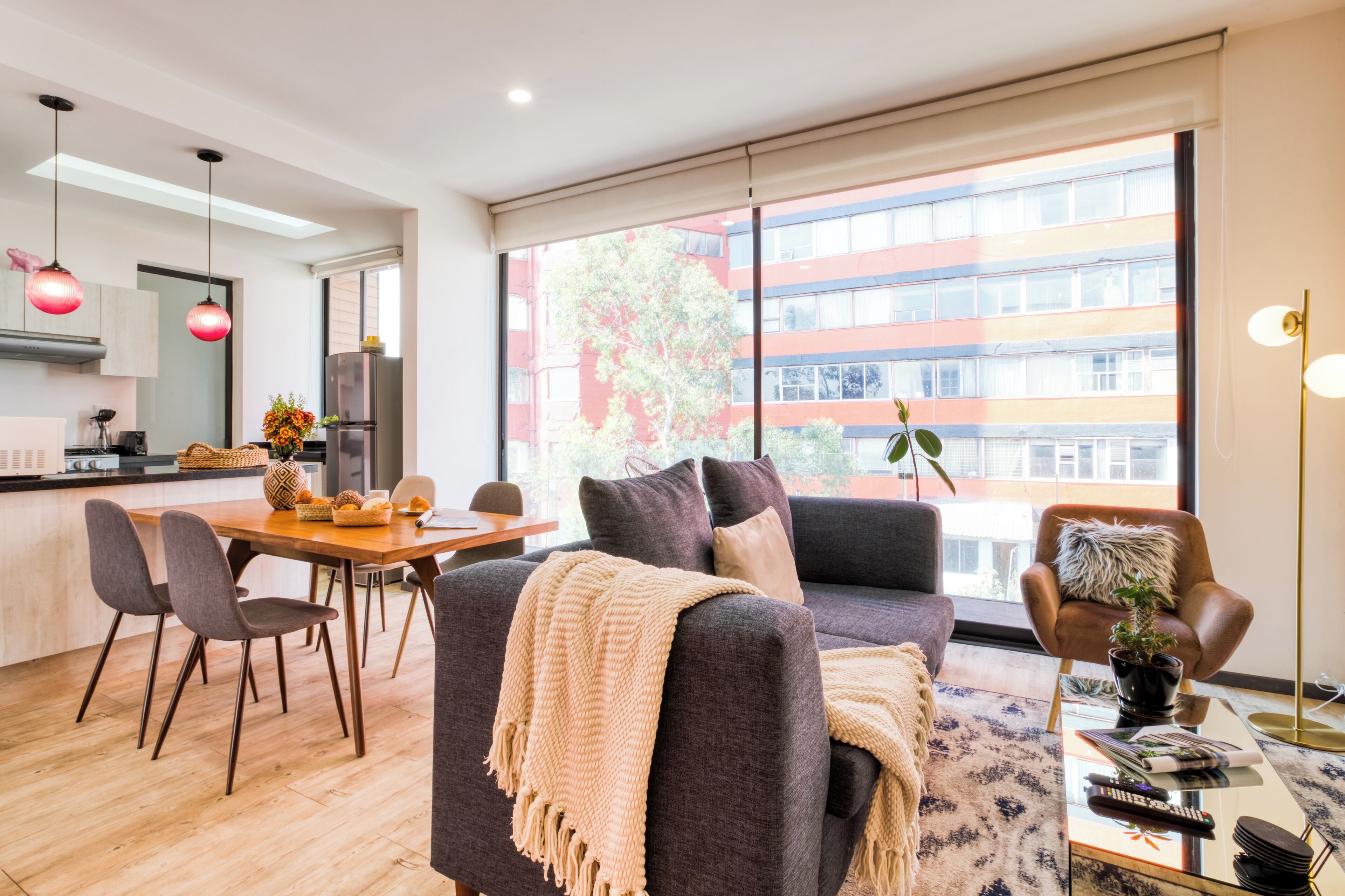 A modern living room with a sofa, dining table, and chairs. There is a large window with a view of buildings outside.