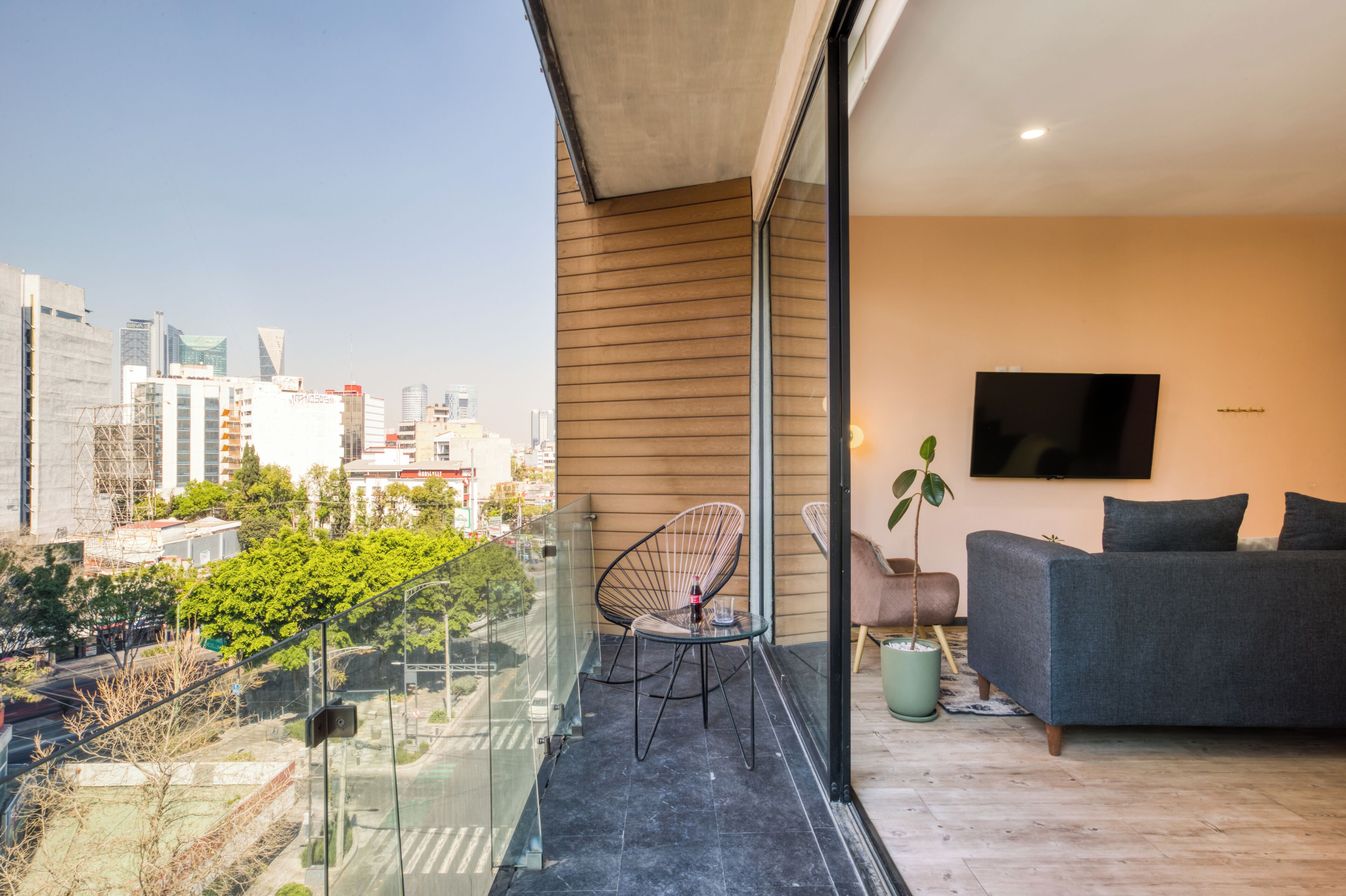 A modern living room with a city view, a glass balcony, a sofa, and a TV mounted on the wall.