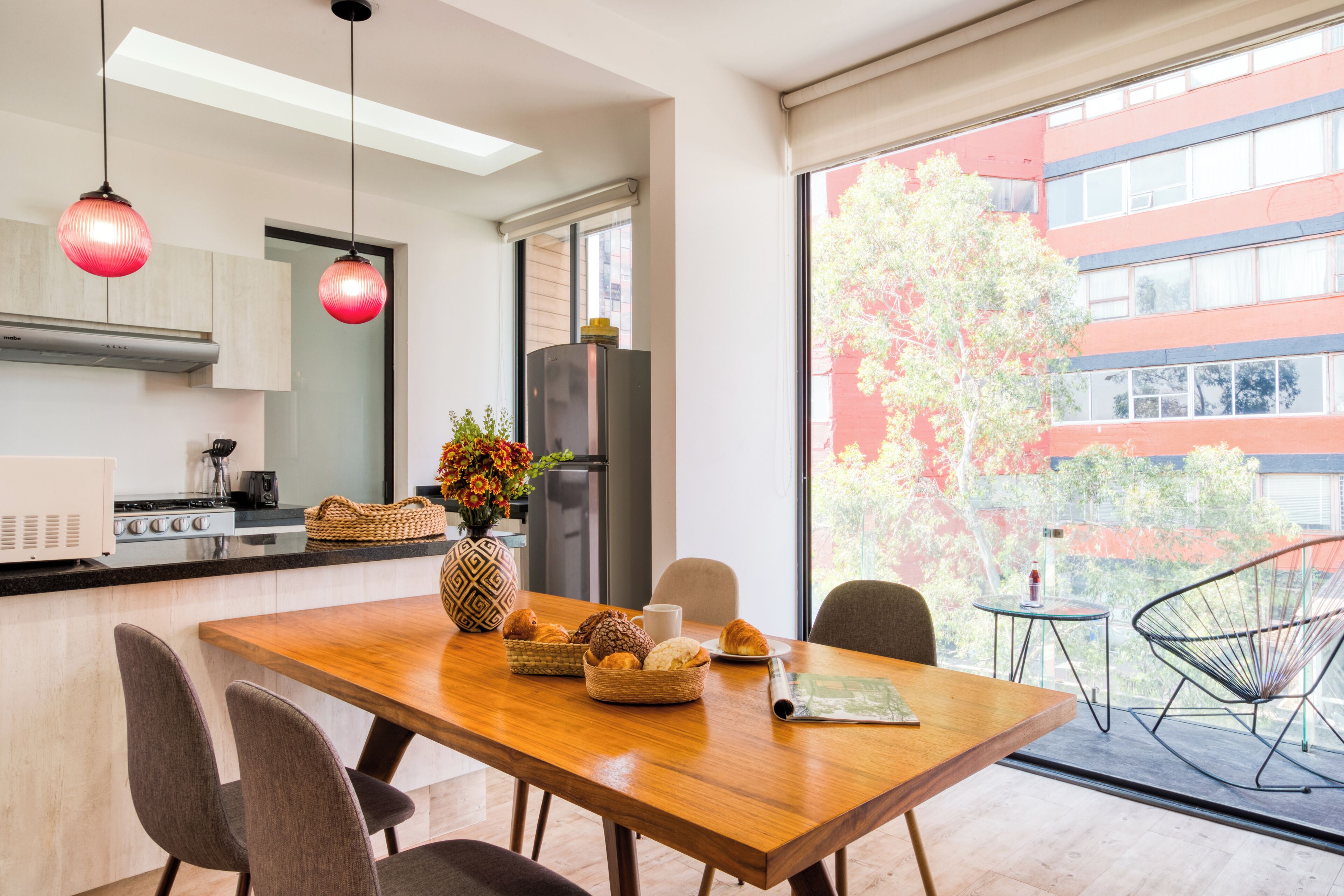 A modern kitchen with a wooden dining table, chairs, and a refrigerator. There is a window with a view of buildings and trees.