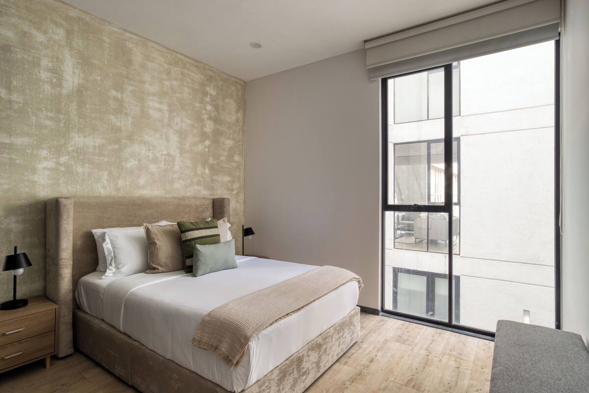 Ein Schlafzimmer mit einem großen Bett, einem Nachttisch, einer Lampe und Blick ins Freie durch ein großes Fenster.