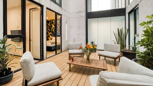 Un área de estar al aire libre con un diseño moderno, que incluye una mesa de madera, sillas acolchadas y plantas en macetas.