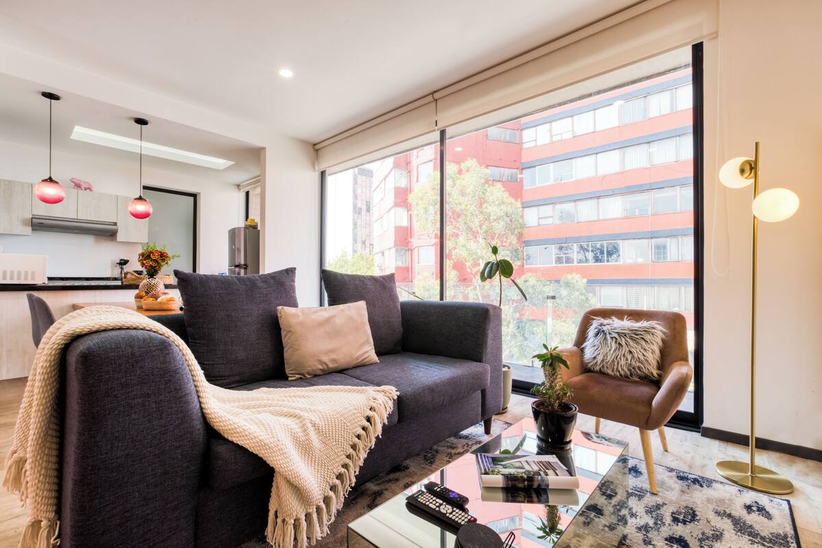Ein modernes Wohnzimmer mit einem dunkelgrauen Sofa, einer beigen Tagesdecke und einem Couchtisch mit einer Zeitschrift und Fernbedienungen. Es gibt einen braunen Sessel mit einem flauschigen Kissen und eine Stehlampe. Ein großes Fenster bietet Blick auf ein rotes Gebäude und Bäume vor der Kulisse.
