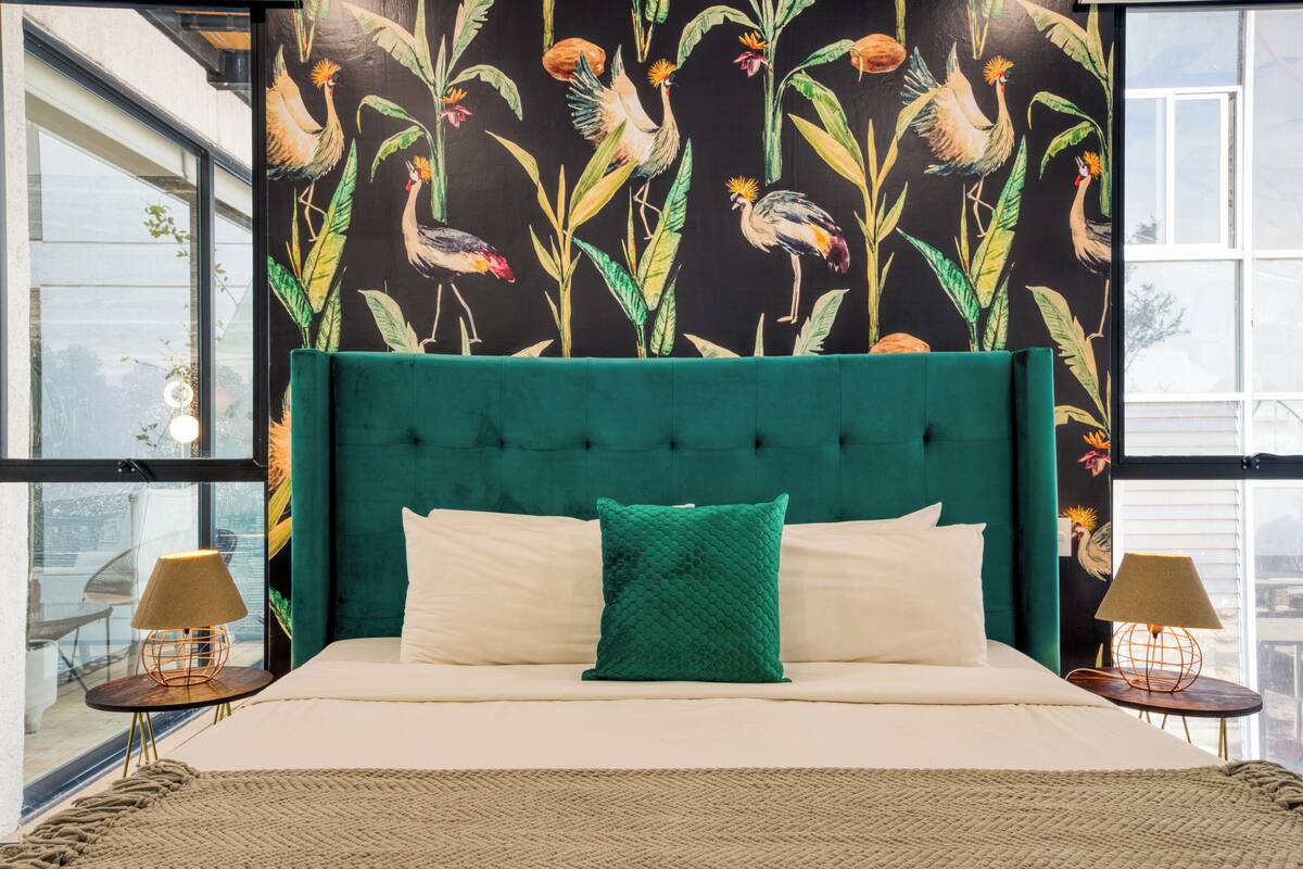 Ein Schlafzimmer mit einem petrolfarbenen, gepolsterten Kopfteil, einem großen Bett mit weißen und grünen Kissen und einem Wandgemälde von Vögeln und Pflanzen.