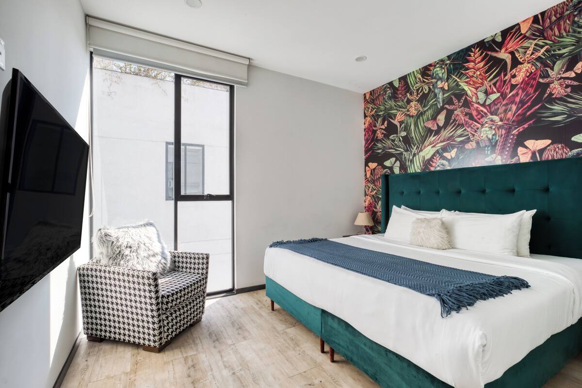 Ein Schlafzimmer mit einem Bett, einem Hundszahn-Sessel, einem Fernseher und einem großen Fenster.