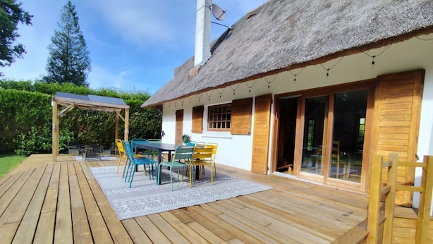 Una terraza de madera con mesa y sillas, una pérgola y una casa con techo de paja y puertas correderas de vidrio.