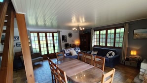 Un comedor con mesa y sillas de madera, un sofá y una araña. Hay ventanas con cortinas, una chimenea y una pared de piedra.