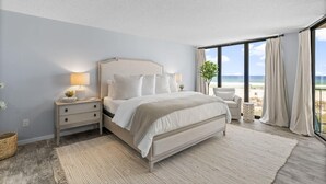 Une chambre avec un grand lit, une table de chevet avec une lampe, une vue sur la plage et une grande fenêtre avec des rideaux.